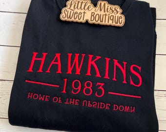 May include: Black sweatshirt with red embroidered text reading "HAWKINS 1983" and "HOME OF THE UPSIDE DOWN". A wooden tag with the text "Little Miss Sweet Boutique" is attached to the sweatshirt.