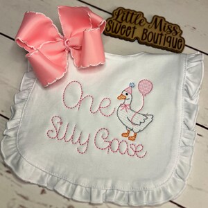 May include: White baby bib with pink embroidered text reading "One Silly Goose." The bib features a cartoon goose wearing a party hat and holding a balloon. A large pink bow is attached to the top left corner. The bib has a ruffled edge.