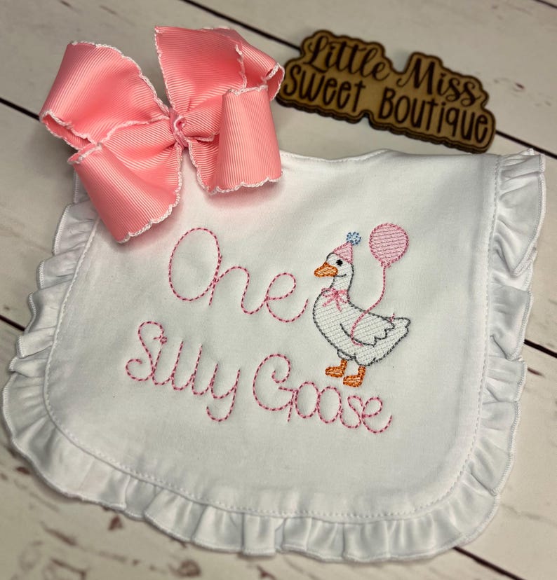 May include: White baby bib with pink embroidered text reading "One Silly Goose." The bib features a cartoon goose wearing a party hat and holding a balloon. A large pink bow is attached to the top left corner. The bib has a ruffled edge.