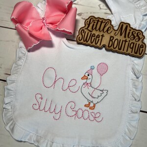 May include: White baby bib with pink ruffle trim and a large pink bow. The bib features embroidered text and a cartoon goose holding a balloon. The text reads "One Silly Goose". A wooden sign says "Little Miss Sweet Boutique".