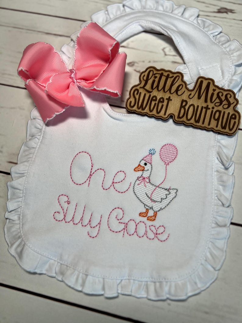 May include: White baby bib with pink ruffle trim and a large pink bow. The bib features embroidered text and a cartoon goose holding a balloon. The text reads "One Silly Goose". A wooden sign says "Little Miss Sweet Boutique".