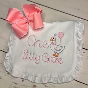 May include: White baby bib with pink embroidered text reading "One Silly Goose" and a cartoon goose wearing a party hat and holding a balloon. A large pink bow is attached to the bib.