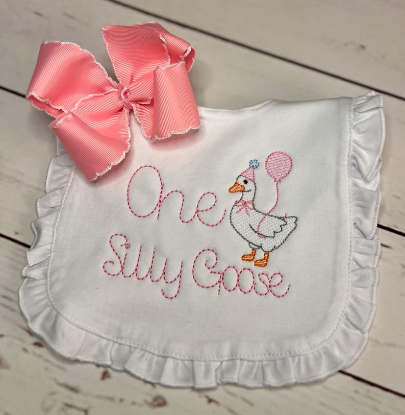 May include: White baby bib with pink embroidered text reading "One Silly Goose" and a cartoon goose wearing a party hat and holding a balloon. A large pink bow is attached to the bib.