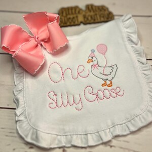 May include: White baby bib with pink embroidered text and a cartoon goose wearing a party hat and holding a balloon. The bib has a ruffled edge and a large pink bow. The text reads "One Silly Goose".