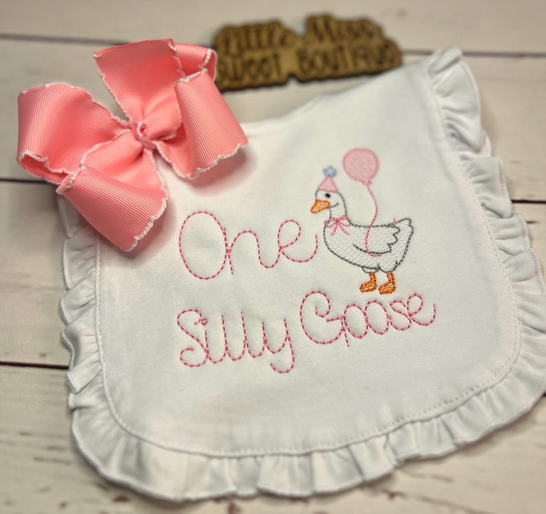 May include: White baby bib with pink embroidered text and a cartoon goose wearing a party hat and holding a balloon. The bib has a ruffled edge and a large pink bow. The text reads "One Silly Goose".