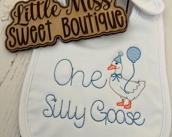 May include: White baby bib with blue embroidered text that reads "One Silly Goose" and a cartoon goose wearing a party hat and holding a balloon. A wooden sign that reads "Little Miss Sweet Boutique" is on top of the bib.