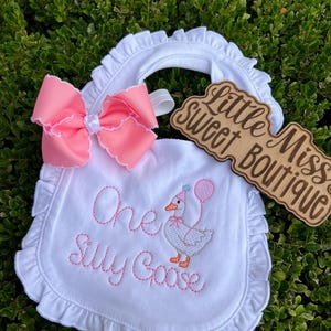 May include: White baby bib with a pink bow and ruffled edges. The bib features embroidered text that reads "One Silly Goose" and an embroidered goose holding a balloon. A wooden sign with the text "Little Miss Sweet Boutique" is also present.