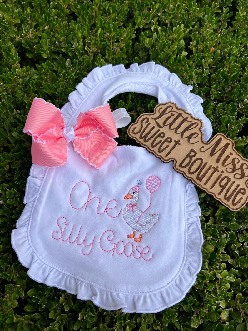 May include: White baby bib with a pink bow and ruffled edges. The bib features embroidered text that reads "One Silly Goose" and an embroidered goose holding a balloon. A wooden sign with the text "Little Miss Sweet Boutique" is also present.