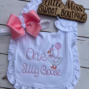 May include: White baby bib with pink embroidered text reading "One Silly Goose" and a cartoon goose wearing a party hat and holding a balloon. A large pink bow with white trim is attached. The bib has a ruffled edge and the text "Little Miss Sweet Boutique" is on a wooden sign.