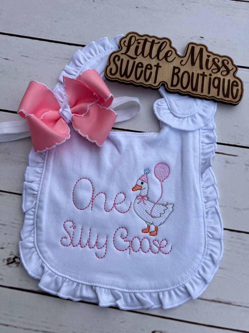 May include: White baby bib with pink embroidered text reading "One Silly Goose" and a cartoon goose wearing a party hat and holding a balloon. A large pink bow with white trim is attached. The bib has a ruffled edge and the text "Little Miss Sweet Boutique" is on a wooden sign.