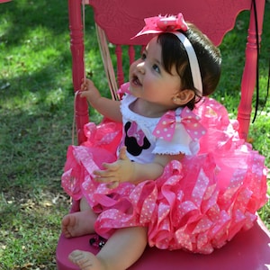 Pode incluir: Uma criança pequena vestindo um vestido rosa e branco de bolinhas com um personagem da Minnie Mouse na frente. O vestido tem uma saia rodada e um laço branco no ombro. A criança está sentada em uma cadeira rosa.