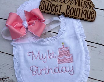 May include: A white baby bib with pink embroidered text reading "My 1st Birthday" and a pink cake design. A matching pink bow headband and a wooden sign that says "Little Miss Sweet Boutique" are also included.