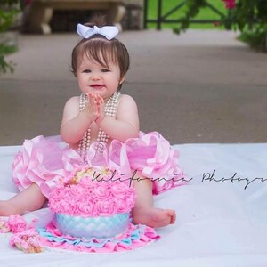 May include: A baby girl in a pink tutu dress with a white bow in her hair sits in front of a pink and blue frosted cake with white and blue rosettes. She is wearing a pearl necklace and has her hands clasped in front of her.
