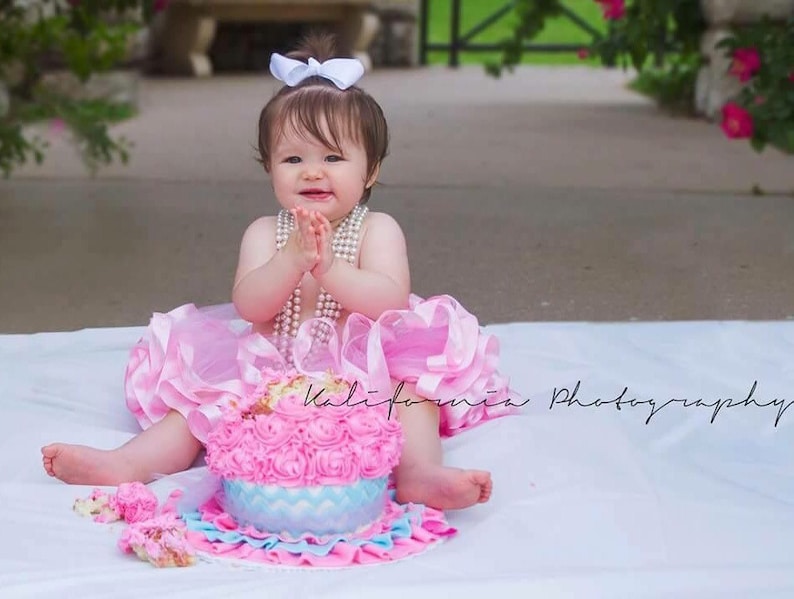 May include: A baby girl in a pink tutu dress with a white bow in her hair sits in front of a pink and blue frosted cake with white and blue rosettes. She is wearing a pearl necklace and has her hands clasped in front of her.