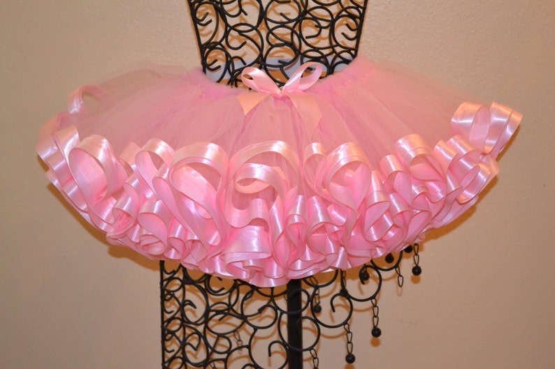 May include: A pink tulle tutu skirt with a pink satin ribbon trim. The ribbon is gathered and ruffled around the bottom of the skirt.