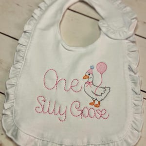 May include: White baby bib with a ruffled edge. The bib features embroidered text that reads "One Silly Goose" in pink thread, along with an embroidered goose wearing a party hat and holding balloons. A cute and festive accessory for a baby.