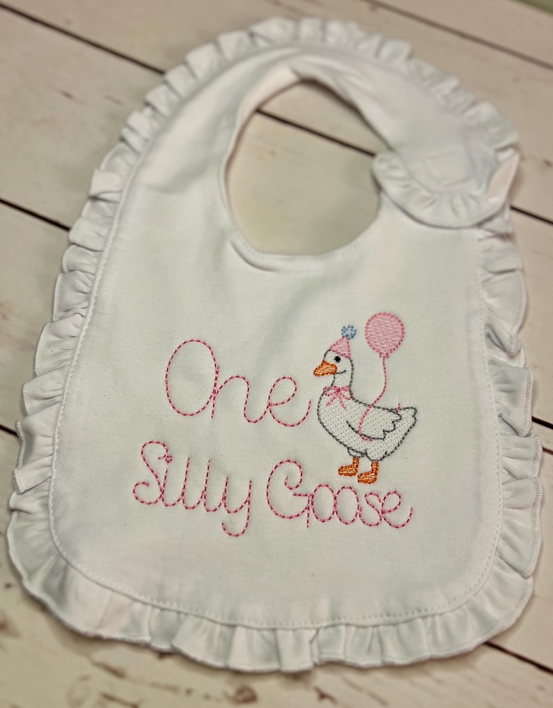 May include: White baby bib with a ruffled edge. The bib features embroidered text that reads "One Silly Goose" in pink thread, along with an embroidered goose wearing a party hat and holding balloons. A cute and festive accessory for a baby.