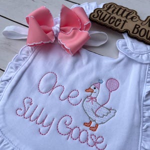 May include: White baby bib with pink embroidered text reading "One Silly Goose" and a cartoon goose holding a balloon. A large pink bow with white trim sits above the bib. The bib has a ruffled edge.