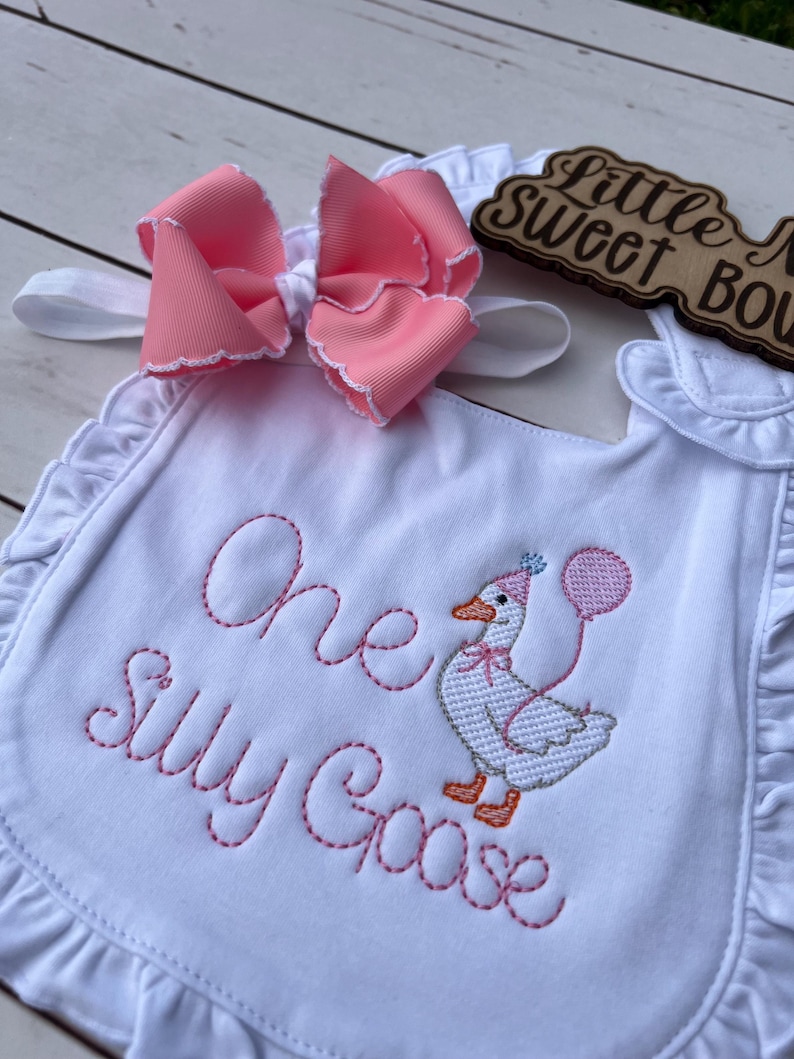 May include: White baby bib with pink embroidered text reading "One Silly Goose" and a cartoon goose holding a balloon. A large pink bow with white trim sits above the bib. The bib has a ruffled edge.