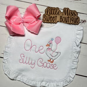 May include: A white baby bib with a ruffled edge, embroidered with the words "One Silly Goose" in pink thread. A cartoon goose wearing a party hat and holding a balloon is also embroidered. A large pink bow is placed above the bib.