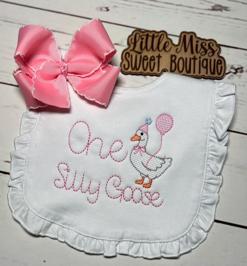May include: A white baby bib with a ruffled edge, embroidered with the words "One Silly Goose" in pink thread. A cartoon goose wearing a party hat and holding a balloon is also embroidered. A large pink bow is placed above the bib.