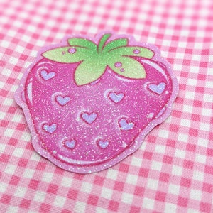 May include: A pink glitter strawberry with a green leafy top and purple hearts. The strawberry is on a pink and white gingham background.