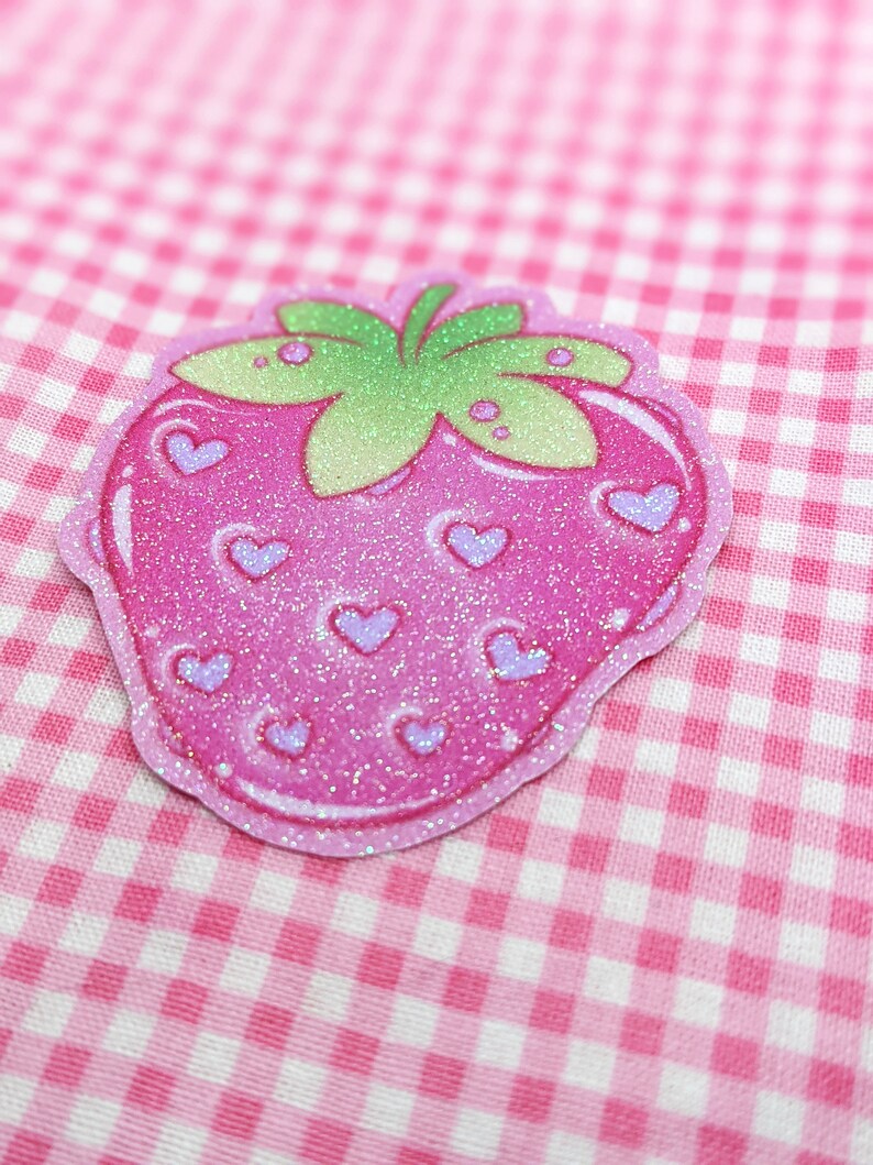 May include: A pink glitter strawberry with a green leafy top and purple hearts. The strawberry is on a pink and white gingham background.