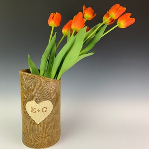 May include: A brown ceramic vase with a wood grain texture holds orange tulips with green stems and leaves. A heart-shaped cutout on the vase displays the initials "E + G". The vase is approximately 8 inches tall.