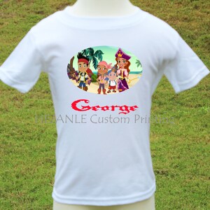 Può includere: T-shirt bianca con una grafica a cartoni animati colorata di personaggi su una spiaggia. La grafica include il testo "George" in rosso, con testo aggiuntivo sottostante. La maglietta è esposta su un manichino su uno sfondo di erba verde.