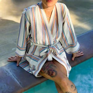 May include: A cream-colored robe featuring vertical stripes in shades of blue, orange, and yellow. The garment has wide sleeves, a V-neckline, and a matching fabric tie belt at the waist. The fabric has a textured weave throughout the entire piece.