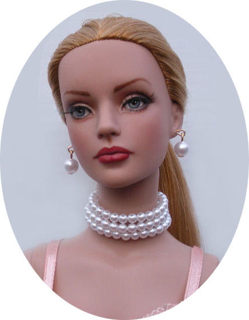 May include: A porcelain doll with blonde hair, wearing a pearl necklace and earrings. The doll is wearing a pink and white dress.