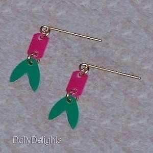 May include: A pair of earrings with pink and green plastic charms. The charms are shaped like a rectangle and a fishtail. The earrings are attached to gold-colored wires.