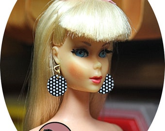 May include: A blonde doll with a pink bow in her hair is wearing black and white polka dot earrings and a black bracelet.