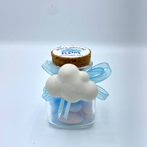 May include: A small glass jar with a cork lid decorated with a white cloud and blue ribbon. The jar contains pink and blue candy and the cork lid has a label that reads "Santa Elena 22 Ottobre 2022".