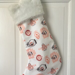 May include: White Christmas stocking with a plush, faux fur cuff. The body of the stocking is patterned with orange and black Astromech Droid graphics and text on a white background. A festive holiday decoration.