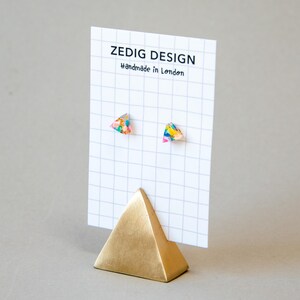 May include: A pair of colorful triangle-shaped stud earrings displayed on a white grid card. The earrings have a vibrant mix of colors. The card is held by a gold pyramid-shaped stand. The card reads "ZEDIG DESIGN Handmade in London."