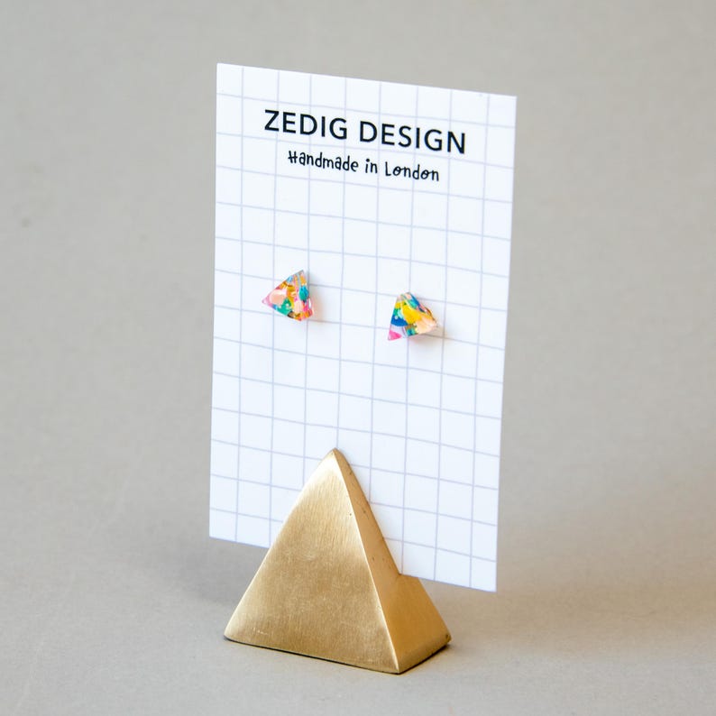 May include: A pair of colorful triangle-shaped stud earrings displayed on a white grid card. The earrings have a vibrant mix of colors. The card is held by a gold pyramid-shaped stand. The card reads "ZEDIG DESIGN Handmade in London."