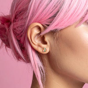 May include: Close-up of a colorful triangular stud earring. The earring features small, colorful stones in shades of pink, blue, and orange. The earring is set against a pink background, with pink hair in the background.