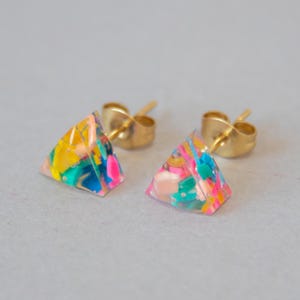 May include: A pair of colorful stud earrings. Each earring is a small, clear pyramid shape with a gold post. The pyramids are filled with a mix of bright colors, including yellow, pink, blue, and green, creating a playful, abstract design.