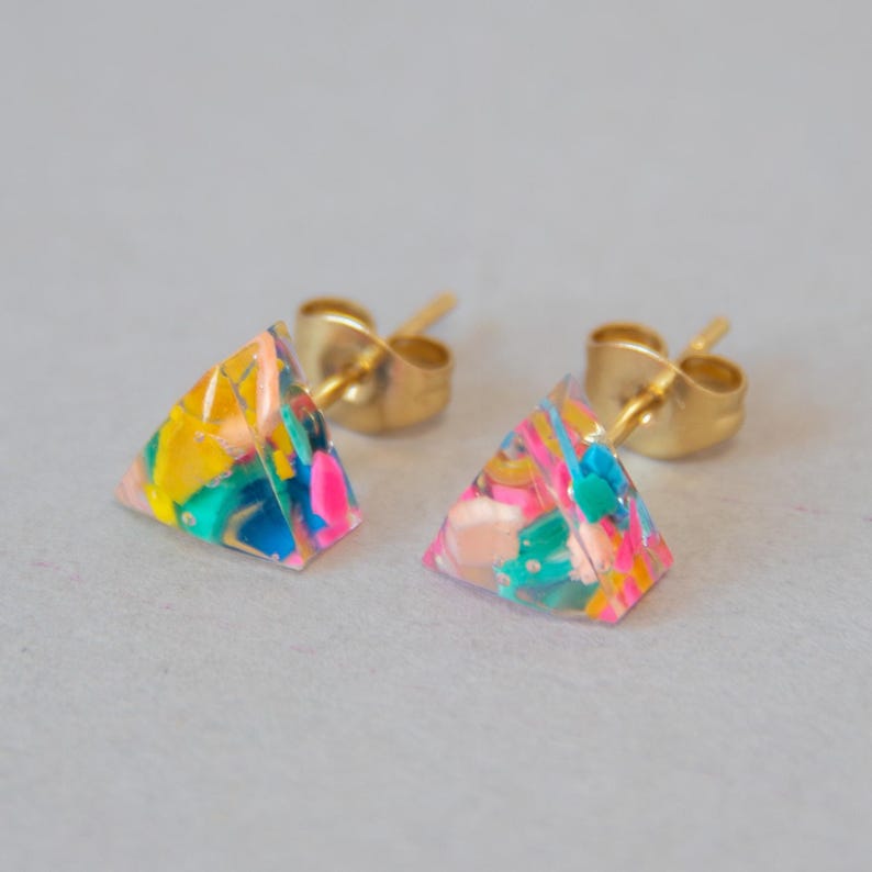 May include: A pair of colorful stud earrings. Each earring is a small, clear pyramid shape with a gold post. The pyramids are filled with a mix of bright colors, including yellow, pink, blue, and green, creating a playful, abstract design.