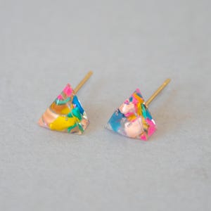 May include: A pair of colorful stud earrings. Each earring is a small, translucent triangle with a gold-colored post. The earrings feature a mix of pink, blue, yellow, and peach-colored elements, creating a vibrant and playful design.