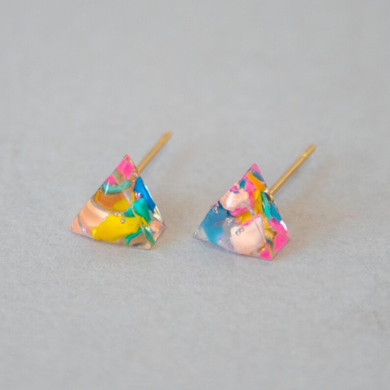 May include: A pair of colorful stud earrings. Each earring is a small, translucent triangle with a gold-colored post. The earrings feature a mix of pink, blue, yellow, and peach-colored elements, creating a vibrant and playful design.