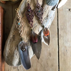 May include: Three macrame earrings with feather details. The earrings have intricate knotwork in brown and burgundy, decorated with beads. Each earring is finished with a natural feather, in shades of grey, brown, and orange.