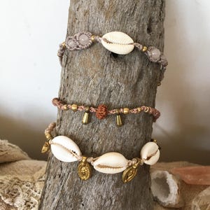 May include: Three handmade macrame bracelets. The top bracelet features a cowrie shell and pink beads. The middle bracelet has small gold beads and charms. The bottom bracelet has cowrie shells and gold charms. The bracelets are displayed on a piece of wood.