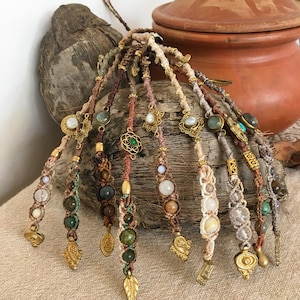 Op de afbeelding: Een verzameling handgemaakte haarwraps met ingewikkelde macraméknopen. De wraps bevatten diverse kralen, waaronder groene, witte en bruine stenen, en zijn versierd met goudkleurige metalen bedels en bladhangers. Het kleurenpalet omvat aardetinten van bruin, beige en groen.