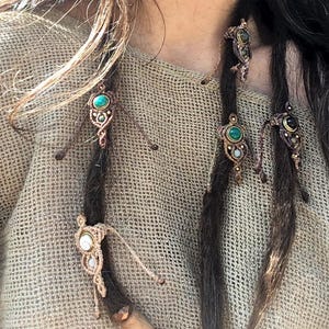 May include: Close-up of several macrame hair accessories with turquoise, green, and white gemstones. The accessories are attached to dark brown dreadlocks. The macrame is a light brown color. The background is a tan, woven fabric.