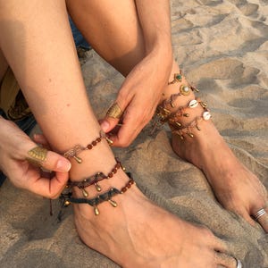 May include: Three layered anklets with decorative beads and charms. The anklets are made of brown, red, and blue thread. The charms are small, gold-coloured teardrop shapes.