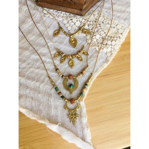 May include: A layered necklace set featuring four gold-toned chains. The necklaces include various gold-toned leaf-shaped charms, small wooden beads, a central circular gold-toned pendant with a small turquoise stone, and a large ornate filigree pendant.