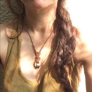 May include: A handmade macrame necklace with a large, oval-shaped pendant. The pendant is a light beige color with brown and orange accents. The necklace is worn over a mustard-colored tank top. A gold-colored nose ring is visible.