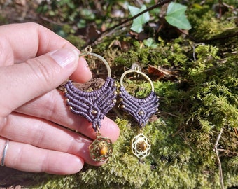 May include: A pair of purple macrame earrings with gold hoops and a tiger's eye stone pendant. The earrings are hanging from a hand.